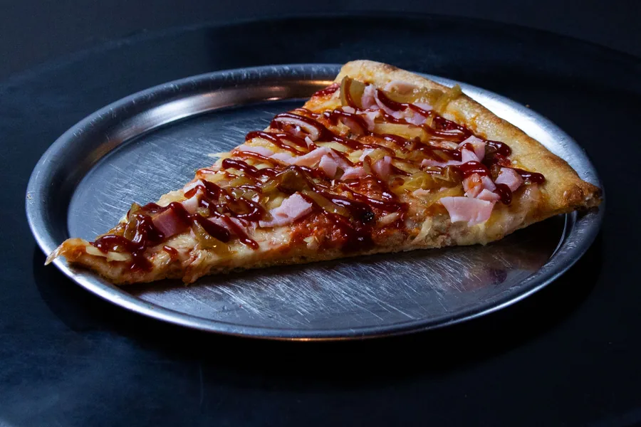 Porción de pizza con jamón, cebolla caramelizada e hilos de salsa barbacoa