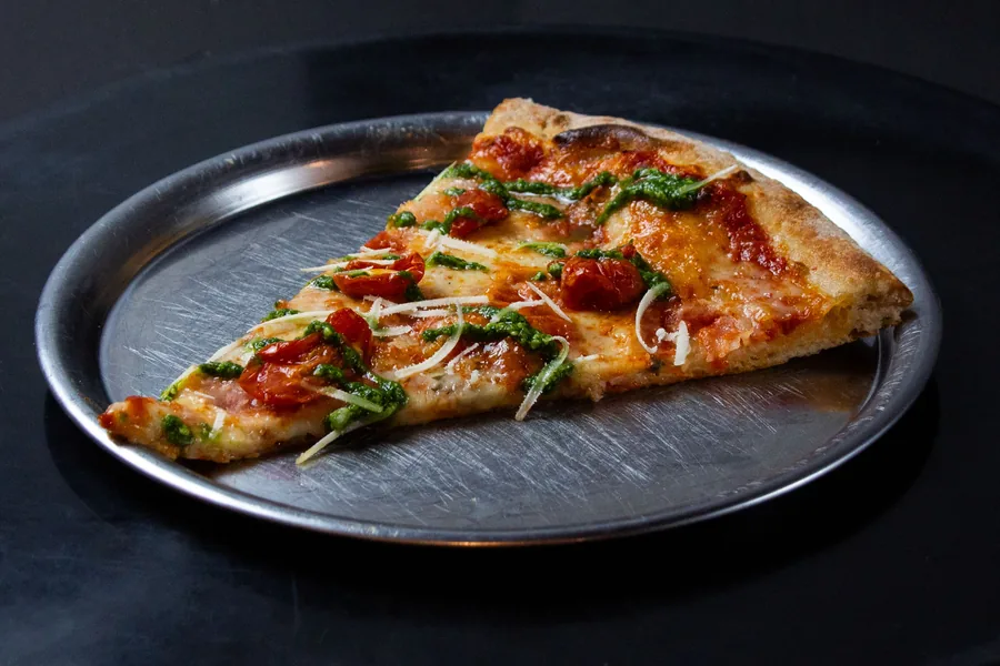 Porción de pizza con tomates cherry, pesto y queso rallado sobre un plato metálico